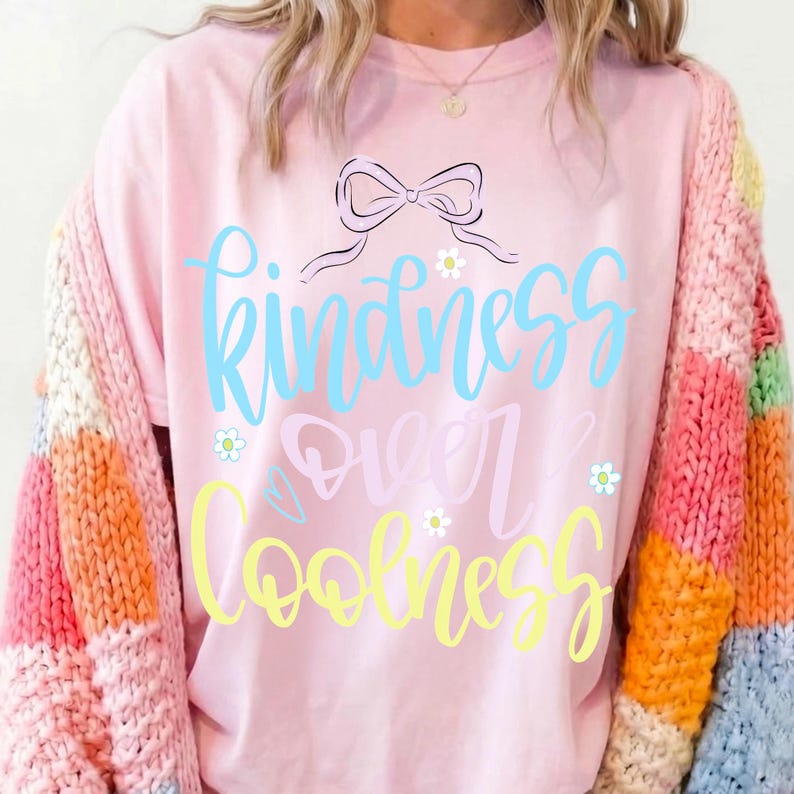 May include: A light pink t-shirt with the words "Kindness Over Coolness" in blue and yellow script. The shirt features a bow and small flower graphics. The person is wearing a colorful, patchwork sweater.