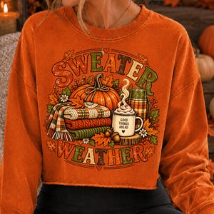Puede incluir: Una sudadera naranja de manga larga con un estampado gr&aacute;fico de una calabaza, una pila de mantas de punto dobladas, un termo y una taza con vapor. El texto dice "SWEATER WEATHER" y "GOOD THINGS AHEAD" con peque&ntilde;os detalles de corazones y hojas.