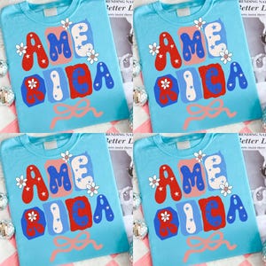 May include: Four light blue t-shirts, each featuring the word "AMERICA" in red, white, and blue, adorned with white flowers and a pink bow. The design is repeated on each shirt.