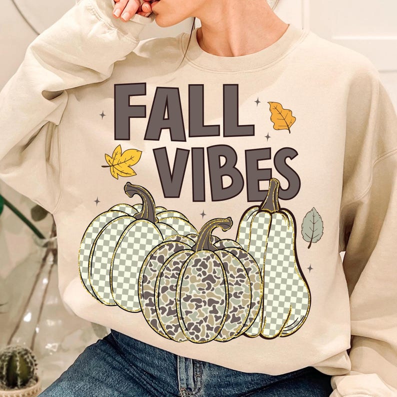 Puede incluir: Una sudadera de manga larga color crema con el texto FALL VIBES en letras marr&oacute;n oscuro. El dise&ntilde;o incluye tres calabazas con patrones de cuadros y camuflaje, acentuadas con peque&ntilde;as ilustraciones de hojas amarillas y verdes y estrellas.