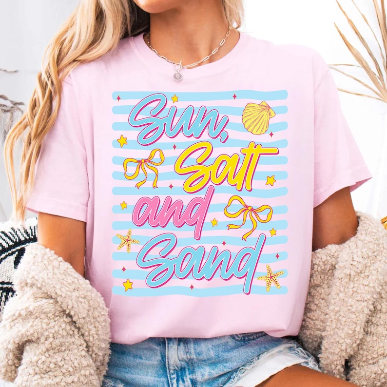 Peut inclure: T-shirt rose clair avec l'inscription "Sun, Salt and Sand" dans un design color&eacute;. Le texte est en bleu, jaune et rose, avec des graphiques de coquillages et d'&eacute;toiles de mer. Le t-shirt a des rayures horizontales bleues.