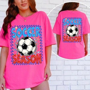 Puede incluir: Camiseta rosa brillante con las palabras "SOCCER SEASON" en azul y rojo, rodeando un gr&aacute;fico de bal&oacute;n de f&uacute;tbol en blanco y negro. El dise&ntilde;o est&aacute; en la parte delantera y trasera de la camiseta. La camiseta tiene mangas cortas y un corte holgado.