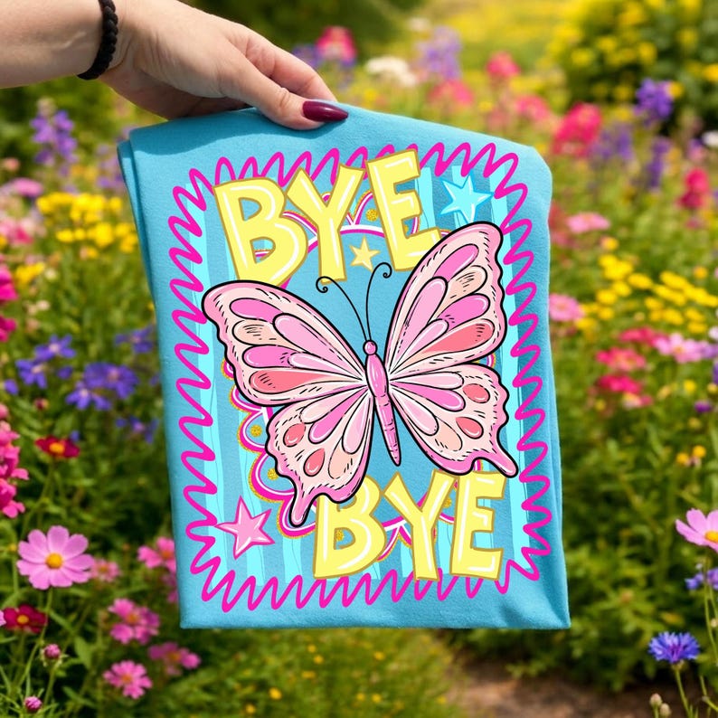 May include: A turquoise t-shirt with a pink butterfly and the words "BYE BYE" in yellow. The design includes pink zig-zag lines and stars. The shirt is held up in front of a colourful flower garden.