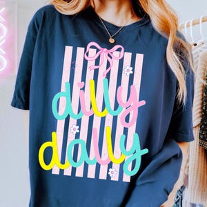 May include: Navy blue t-shirt featuring the words "dilly dally" in a vibrant, whimsical font. The design incorporates pink vertical stripes, a pink bow, and floral details. A relaxed, comfortable tee.