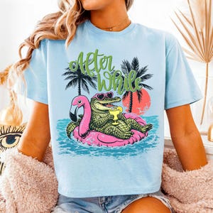 Pu&ograve; includere: T-shirt azzurra con una grafica raffigurante un alligatore con occhiali da sole e un drink, che galleggia su un galleggiante a forma di fenicottero rosa. Il testo "After While" &egrave; sopra l'alligatore. Palme e un tramonto sullo sfondo.