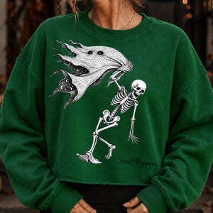 K&ouml;nnte beinhalten: Ein dunkelgr&uuml;nes Langarm-Sweatshirt mit einem wei&szlig;en Grafikdruck eines Skeletts, das ein geisterhaftes Laken mit Sternenhimmel-Muster h&auml;lt. Der Text "Sheet Happens.." ist in schwarzer Schrift unten rechts aufgedruckt.