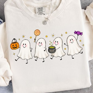 May include: A cream-colored crewneck sweatshirt featuring a graphic of four white ghosts. The ghosts are holding a jack-o'-lantern bucket, an orange lollipop, a black cauldron with green liquid, and a purple candy. Small yellow stars surround them.