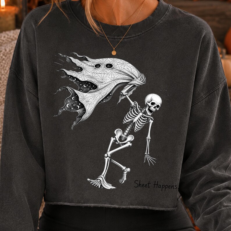 K&ouml;nnte beinhalten: Ein anthrazitfarbenes, bauchfreies Sweatshirt mit einem wei&szlig;en Grafikdruck eines Skeletts, das ein flie&szlig;endes Laken h&auml;lt, welches ein Sternenhimmel-Muster enth&uuml;llt. Der Text "Sheet Happens" steht in schwarzer Schreibschrift unten rechts.