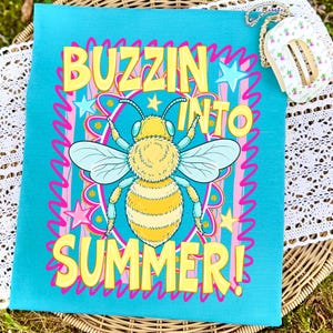 K&ouml;nnte beinhalten: Ein t&uuml;rkisfarbener Stoff mit einer gro&szlig;en gelben Bienenillustration und den Worten "BUZZIN INTO SUMMER!" in gelben Buchstaben. Die Biene hat hellblaue Fl&uuml;gel und ist von rosa und blauen Sternen und wellenf&ouml;rmigen Linien umgeben. Eine kleine florale Geldb&ouml;rse ist im Hintergrund.