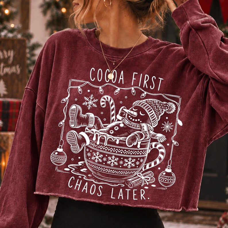 Op de afbeelding: Een bordeauxrood sweatshirt met lange mouwen en een rauwe zoom, voorzien van een witte afbeelding van een peperkoekmannetje in een mok. De tekst luidt COCOA FIRST CHAOS LATER. Het ontwerp bevat sneeuwvlokken, kerstverlichting en ornamenten.