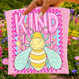 May include: Pink t-shirt with a cartoon bee illustration and the word "KIND" in large letters. The bee is yellow and white with blue wings, surrounded by stars and a wavy pink border. The background is a colorful garden.