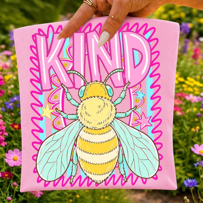 May include: Pink t-shirt with a cartoon bee illustration and the word "KIND" in large letters. The bee is yellow and white with blue wings, surrounded by stars and a wavy pink border. The background is a colorful garden.