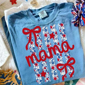 Op de afbeelding: Een lichtblauw t-shirt met het woord "mama" in rode letters. Het shirt heeft verticale strepen met een bloemenpatroon in rood, wit en blauw, samen met rode strikken en sterren. Het shirt is maat XL 16-66.