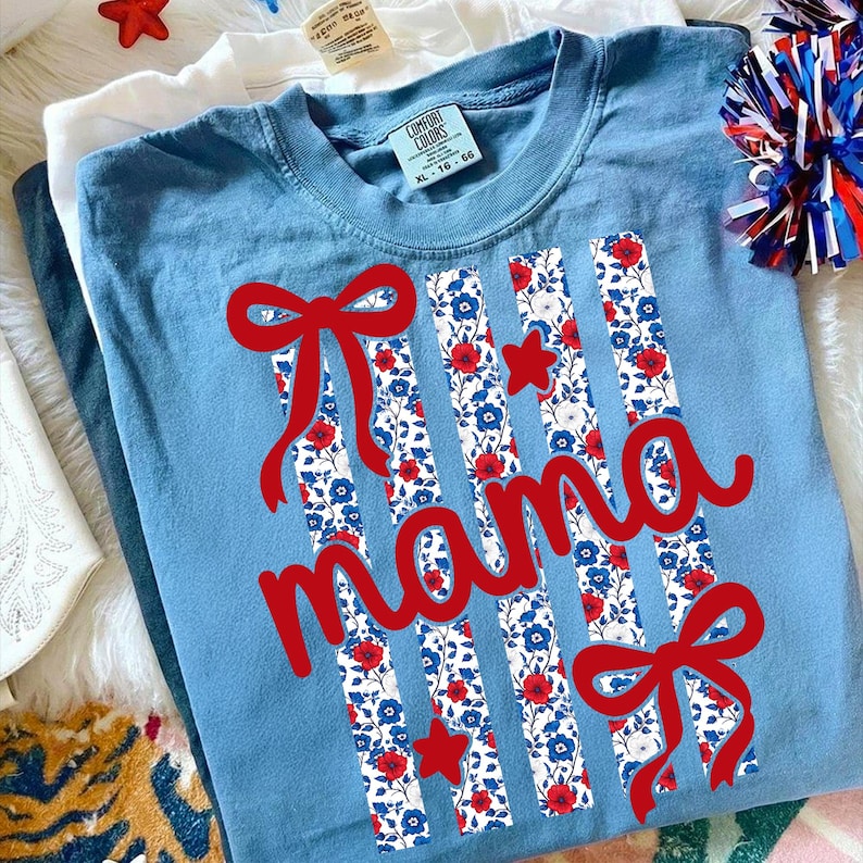 Op de afbeelding: Een lichtblauw t-shirt met het woord "mama" in rode letters. Het shirt heeft verticale strepen met een bloemenpatroon in rood, wit en blauw, samen met rode strikken en sterren. Het shirt is maat XL 16-66.