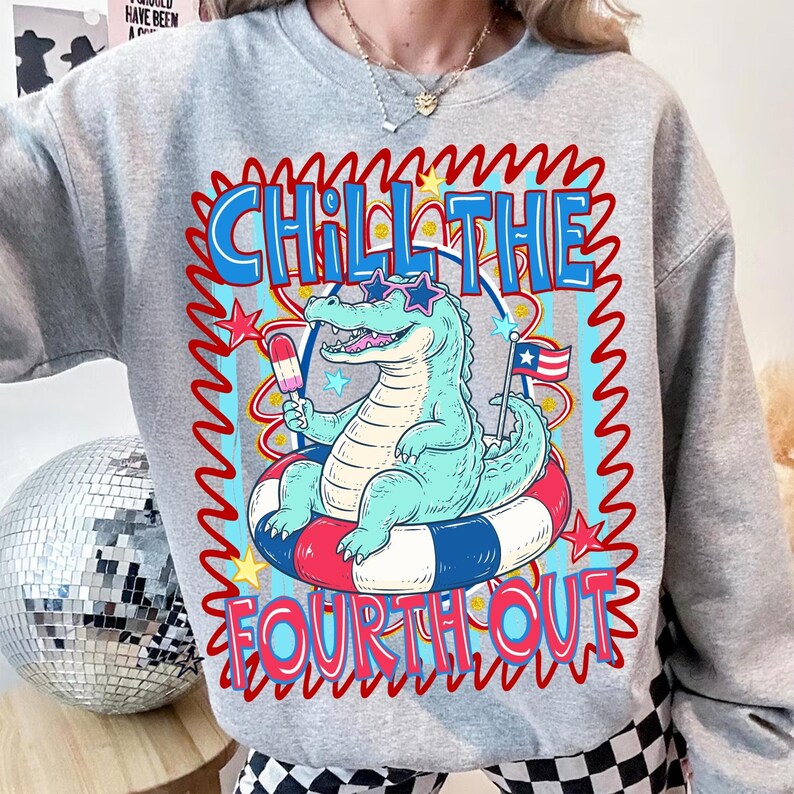 Puede incluir: Una sudadera gris con un cocodrilo de dibujos animados con gafas de sol y flotando en un flotador inflable rojo, blanco y azul. El texto "CHILL THE FOURTH OUT" se muestra en una fuente colorida y ondulada, con estrellas y un helado.