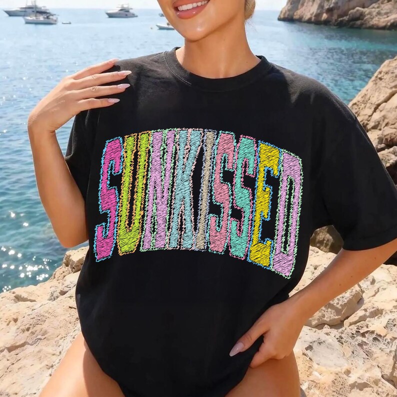 May include: Black t-shirt featuring the word "SUNKISSED" embroidered in a vibrant, multi-coloured design. The text uses shades of pink, yellow, blue, and green. The shirt has a relaxed fit.