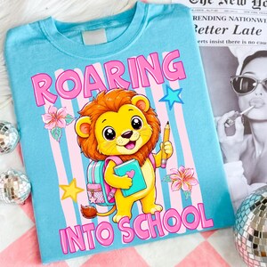 May include: A light blue t-shirt featuring a cartoon lion holding a pencil and a book. The graphic includes pink and blue vertical stripes, pink flowers, yellow stars, and the text ROARING INTO SCHOOL in bold pink letters across the top and bottom.