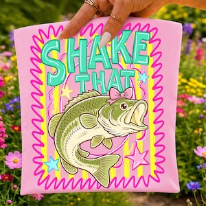 Op de afbeelding: Roze t-shirt met een cartoon bas met een roze strik en de woorden "SHAKE THAT" in turquoise en geel. Het ontwerp bevat gele strepen, roze zigzags en steraccenten. Het t-shirt wordt door een persoon vastgehouden.