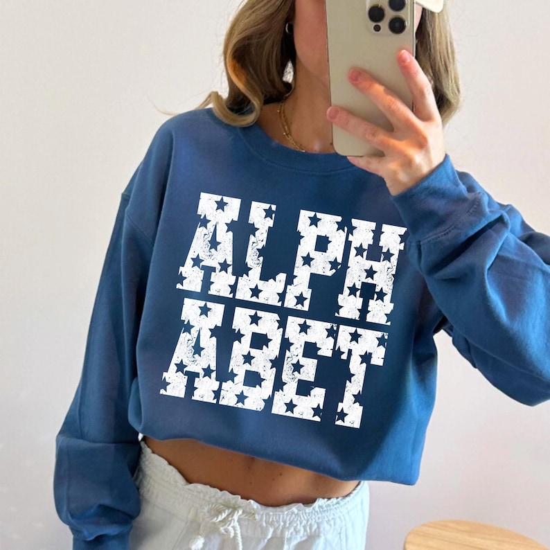 May include: A cropped, navy blue sweatshirt with the text "ALPHABET" in white, star-filled letters. The long-sleeved top is a casual style, suitable for everyday wear.