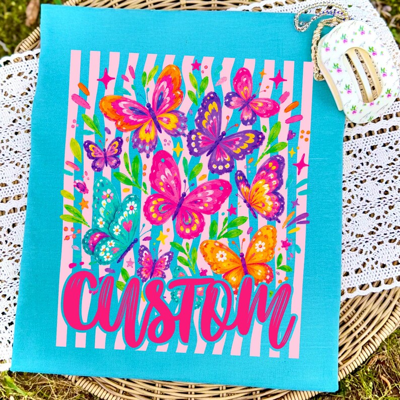 May include: A bright turquoise fabric featuring a central graphic of multiple colourful butterflies in shades of pink, orange, purple, and teal, set against a background of vertical light pink stripes. The word CUSTOM is printed in bold pink script.