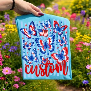 Peut inclure: Un t-shirt bleu clair orn&eacute; d'un motif de papillons aux ailes rouges, blanches et bleues, d'un petit drapeau am&eacute;ricain et du mot custom &eacute;crit en lettres cursives rouges. Le design inclut des rayures verticales bleu clair et des &eacute;toiles rouges.
