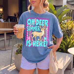 K&ouml;nnte beinhalten: Blaues T-Shirt mit dem Text "OH DEER SUMMER IS HERE!" und einer Hirschgrafik. Das Shirt wird mit gestreiften Shorts kombiniert. Das Bild zeigt auch eine beige Handtasche und ein Getr&auml;nk in einem durchsichtigen Becher.