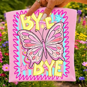 May include: Pink graphic tee featuring a butterfly design. The butterfly is pink with darker pink and peach accents. The words "BYE BYE" are in yellow, with a pink wavy border and blue stripes. Stars are also included in the design.