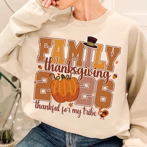 Pode incluir: Uma camisola bege de gola redonda com um gr&aacute;fico que inclui o texto FAMILY thanksgiving 2026 e thankful for my tribe. O design apresenta uma ab&oacute;bora laranja grande, um chap&eacute;u de peregrino castanho, um peru e folhas de outono.