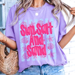 K&ouml;nnte beinhalten: Ein helllila T-Shirt mit dem Schriftzug "Sun Salt And Sand" in leuchtendem Pink. Das Design umfasst hellblaue vertikale Streifen und rosa Muschel- und Seestern-Grafiken. Ein l&auml;ssiges Kleidungsst&uuml;ck zum Thema Strand.