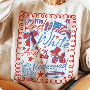 May include: Cream-colored sweatshirt with a red and blue graphic design. The design includes the text "1776 Red White And Redeemed" along with illustrations of an American flag, fireworks, and other patriotic symbols.