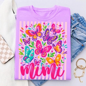 May include: A light purple t-shirt featuring a graphic print of colorful butterflies in shades of pink, yellow, blue, and purple, accented with green leaves and small stars. The word Mimi is printed in a large, bold, bright pink cursive font.