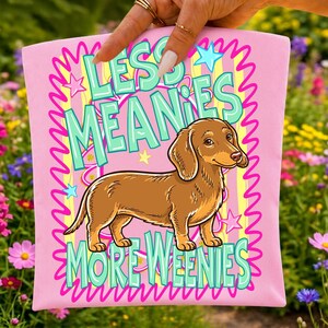 May include: Pink t-shirt with a cartoon dachshund illustration. The shirt features the text "LESS MEANIES MORE WEENIES" in a playful font, with stars and wavy lines. The dachshund is brown with a light brown ear.