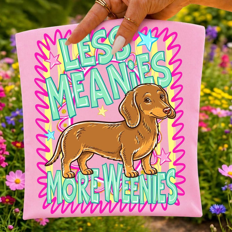 May include: Pink t-shirt with a cartoon dachshund illustration. The shirt features the text "LESS MEANIES MORE WEENIES" in a playful font, with stars and wavy lines. The dachshund is brown with a light brown ear.