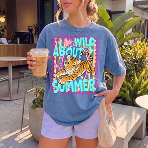 K&ouml;nnte beinhalten: Blaues T-Shirt mit einem farbenfrohen Aufdruck mit der Aufschrift "WILD ABOUT SUMMER" und einem Zeichentiger. Das Shirt wird mit gestreiften Shorts kombiniert. Eine Person h&auml;lt ein Getr&auml;nk in einem durchsichtigen Plastikbecher.