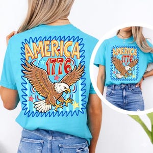 May include: A light blue t-shirt featuring a graphic of a bald eagle with outstretched wings, the word "AMERICA" in yellow and orange, and the number "1776" in red. The design is framed by a blue wavy border and stars.