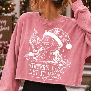May include: A dusty rose long-sleeve cropped sweatshirt featuring a white graphic of a melting snowman wearing a Santa hat and holding a mug. The text reads, "WINTER'S PACE, LET IT MELT." with a small heart icon below the lettering.