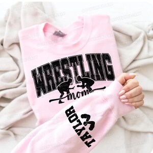 May include: A light pink sweatshirt with the word "WRESTLING" in large, distressed black letters. Below the word are two wrestler silhouettes and the word "mom". The sleeve displays the name "TAYLOR" and wrestling figures.