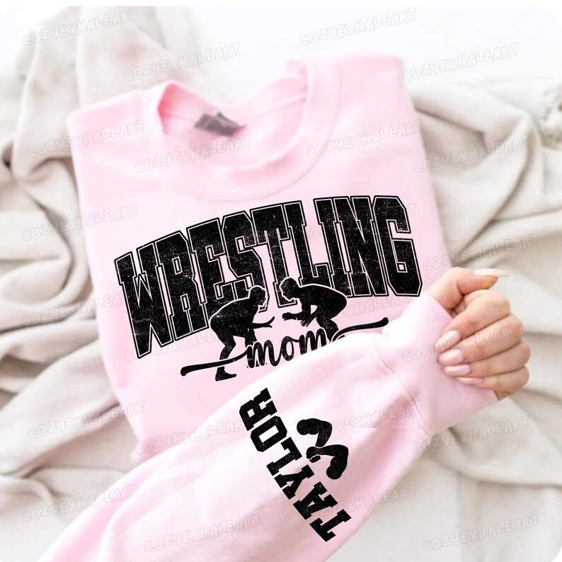 May include: A light pink sweatshirt with the word "WRESTLING" in large, distressed black letters. Below the word are two wrestler silhouettes and the word "mom". The sleeve displays the name "TAYLOR" and wrestling figures.