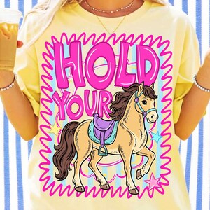 May include: Yellow t-shirt with a graphic of a horse and the text "HOLD YOUR". The horse is tan with a purple saddle and blue bridle. The text is pink with a blue and pink wavy border.