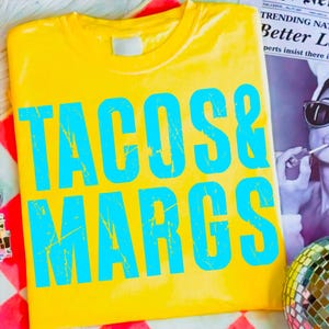 May include: A yellow t-shirt with the words "TACOS & MARGS" in large, distressed, turquoise letters. The shirt is folded and rests on a red and white checkered surface. A newspaper and disco ball are in the background.