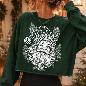 K&ouml;nnte beinhalten: Ein dunkelgr&uuml;nes, lang&auml;rmeliges bauchfreies Sweatshirt mit einem wei&szlig;en Grafikdruck eines zwinkernden Weihnachtsmanns, der eine M&uuml;tze mit Leopardenmuster tr&auml;gt, umgeben von Tannennadeln und Stechpalmenbeeren, mit einem rohen Saum.