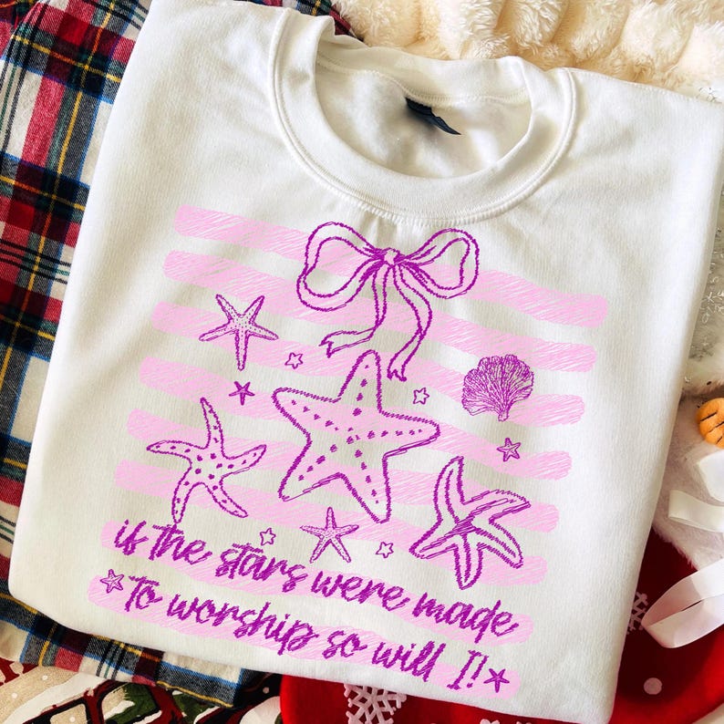 Op de afbeelding: Witte sweatshirt met een roze en paars ontwerp van zeesterren en strikken. De tekst luidt "if the stars were made to worship so will I!" Het ontwerp bevat roze horizontale strepen.