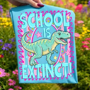 Op de afbeelding: Een lichtblauw t-shirt met de tekst "SCHOOL IS EXTINCT!" in een bubbellettertype. Een cartoon Tyrannosaurus Rex staat in het midden, omringd door sterren en een roze en blauwe rand. Het t-shirt wordt door een persoon vastgehouden.