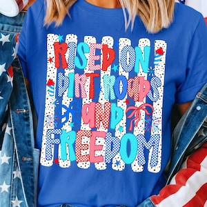 Op de afbeelding: Een koningsblauw T-shirt met korte mouwen en een grafische print van verticale witte strepen met zwarte stippen. De tekst RAISED ON DIRT ROADS AND FREEDOM staat in rood-wit-blauw gepatroonde letters, versierd met vuurwerk en strikjes.