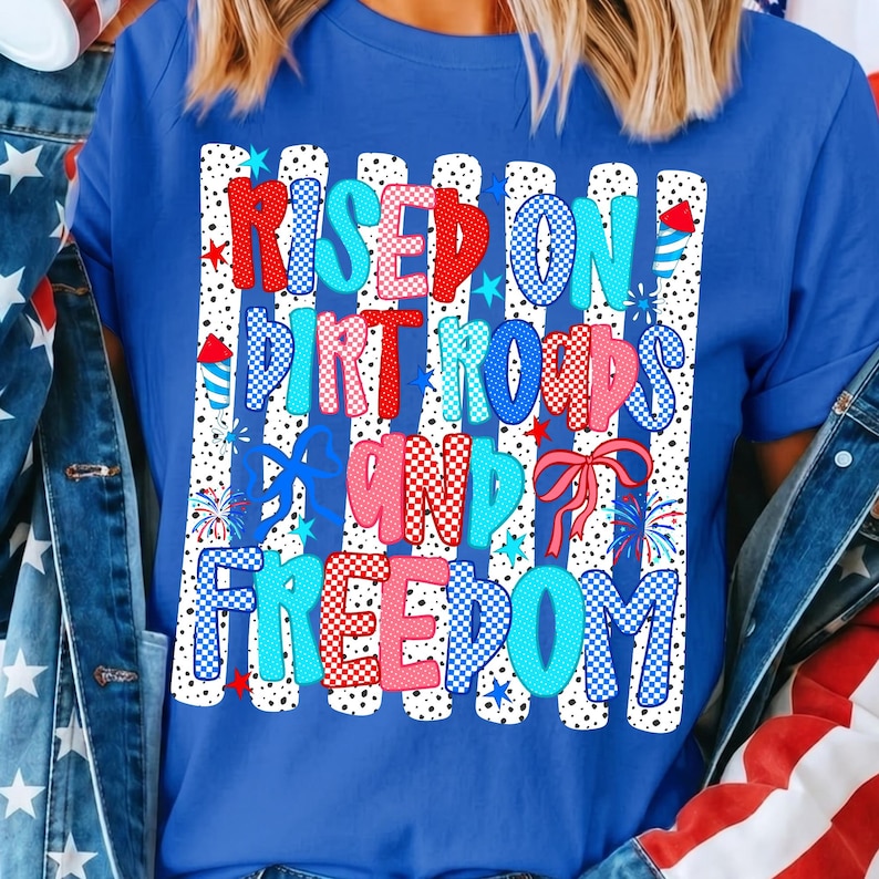 Op de afbeelding: Een koningsblauw T-shirt met korte mouwen en een grafische print van verticale witte strepen met zwarte stippen. De tekst RAISED ON DIRT ROADS AND FREEDOM staat in rood-wit-blauw gepatroonde letters, versierd met vuurwerk en strikjes.