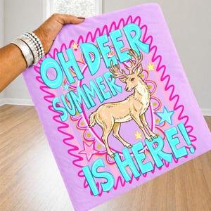 K&ouml;nnte beinhalten: Ein helllila Handtuch mit dem Text "OH DEER SUMMER IS HERE!" in T&uuml;rkis. In der Mitte befindet sich ein Cartoon-Hirsch mit Geweih. Das Design umfasst rosa Sterne und einen rosa Wellenrand.