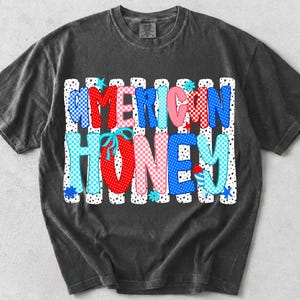 May include: A charcoal grey short-sleeve t-shirt featuring a graphic print with the text AMERICAN HONEY. The letters are filled with red, white, and blue patterns including polka dots and checkers, accented by small stars, flowers, and a firework.