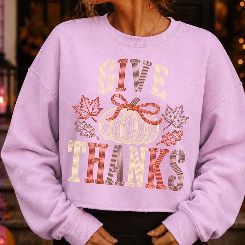 Puede incluir: Una sudadera de manga larga color morado claro con el texto GIVE THANKS en una fuente de rayas multicolor. Centrada entre las palabras hay una calabaza de l&iacute;neas con un lazo rojo, rodeada de ilustraciones de hojas oto&ntilde;ales en rojo y marr&oacute;n.