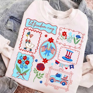 May include: White sweatshirt with patriotic graphics. Features a goose in a bow, American flags, flowers, a top hat, and a truck. The text "let freedom ring" is displayed in a blue banner. The design is in red, white, and blue.