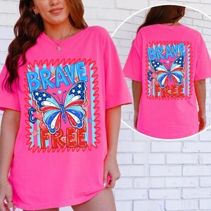 May include: A vibrant pink t-shirt with a butterfly design in red, white, and blue. The words "BRAVE" and "FREE" are printed above and below the butterfly. The same design is on the back of the shirt.